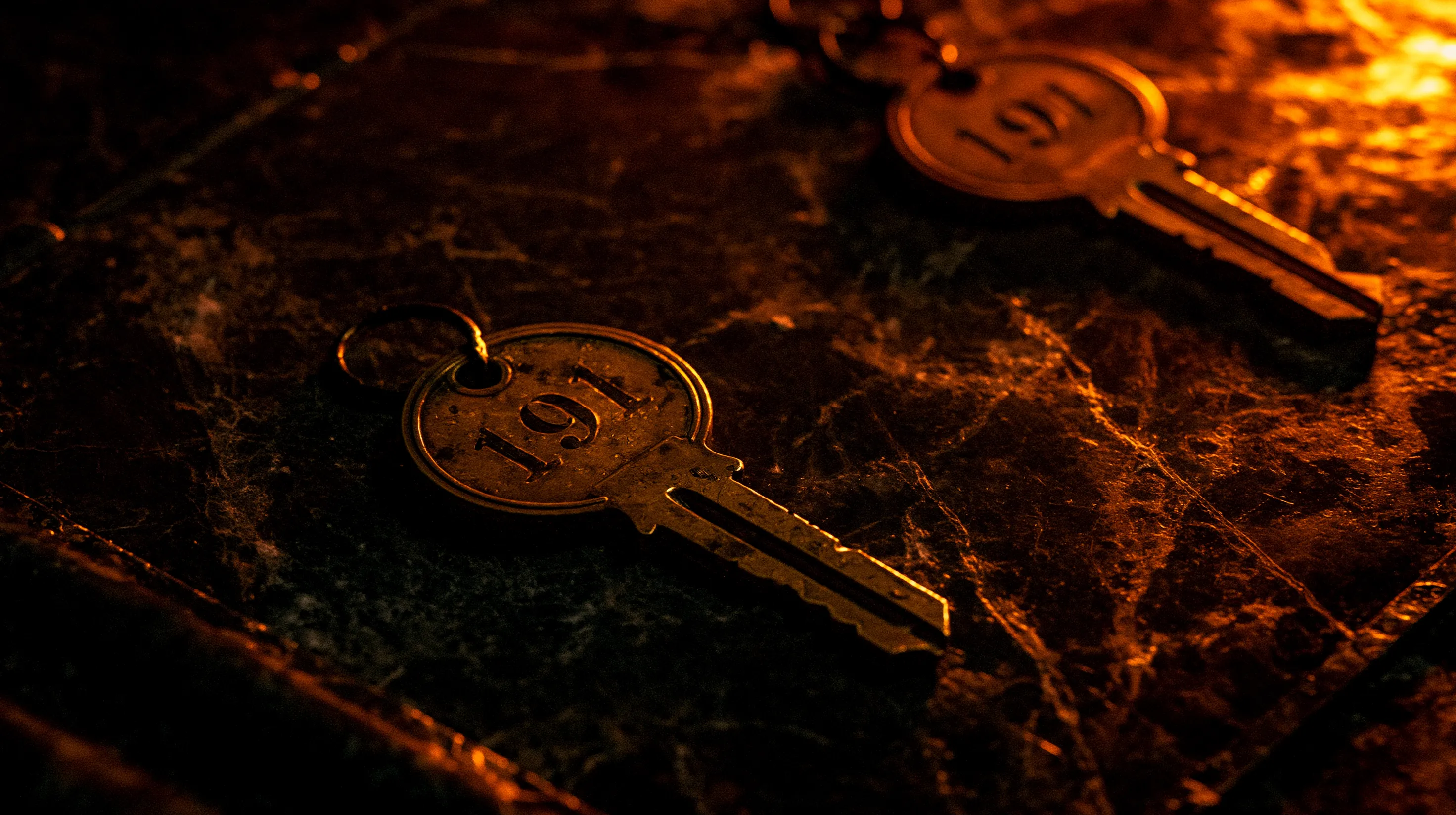 Two identical brass hotel keys stamped 191 on dark gold-veined marble, one lit in a pool of amber light, its duplicate receding into shadow.